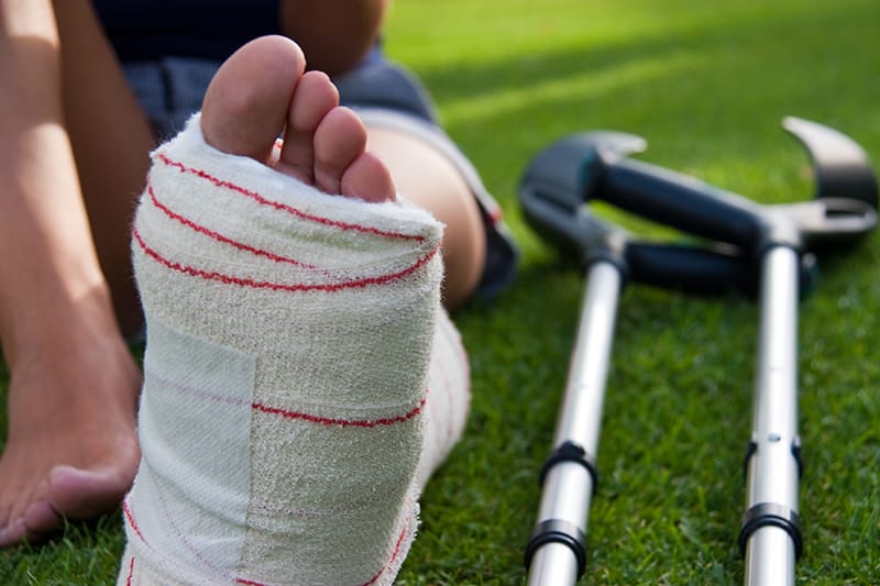 young girl sits on grass with foot in cast, crutches lay by her side